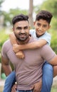 Vertical shot of joyful indian father piggy back his son by looking at camera at park - concept of father love, playful