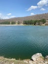 Vertical shot of a lake surrounded by green trees and hillside plants under the blue sky Royalty Free Stock Photo