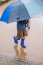 Vertical shot of a little boy with an umbrella and blue rubber boots walking through the wet ground Royalty Free Stock Photo