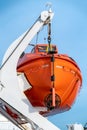 Vertical shot of orange life boat hanging on a crane onboard sailing ocean ship with blue sky in the back. Royalty Free Stock Photo