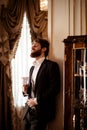 Vertical shot of pleased bearded young businessman wears black formal suit holds glass and drinks beverage, stands in