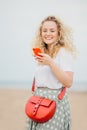 Vertical shot of pleased young female dressed in fashionable clothes, carries bag, holds modern cell phone, texts feedback to frie Royalty Free Stock Photo