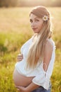 Vertical shot of pregnant female keeps hand on tummy, anticipates baby, has long hair, walks across green field, looks directly at