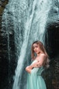 Vertical shot of the pretty woman wearing a long Tiffani dress and standing near the waterfall