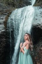 Vertical shot of the pretty woman wearing a long Tiffani dress and standing near the waterfall