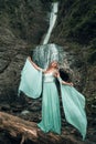 Vertical shot of the pretty woman wearing a long Tiffani dress and standing near the waterfall