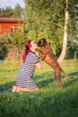 Vertical shot of a redheaded woman kneeling down in a lovely green park to give her boxer a kiss