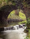 Vertical shot of a small waterfall flowing to a river under the arch of a stone bridge Royalty Free Stock Photo