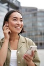 Vertical shot of smiling asian woman in wireless headphones, enjoys listening to music in earphones, holds mobile phone Royalty Free Stock Photo