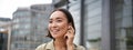 Vertical shot of smiling asian woman in wireless headphones, enjoys listening to music in earphones, holds mobile phone Royalty Free Stock Photo