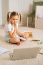 Vertical shot of smiling blonde pretty primary girl child writing homework in copybook sitting on floor at home, looking Royalty Free Stock Photo