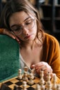 Vertical shot of thinking attarctive young woman wearing elegant eyeglasses making chess move with knight piece sitting Royalty Free Stock Photo