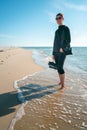 Vertical shot of a woman in dark outdoor clothes with shoes in her right hand, standing barefoot on a beach, with tide