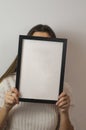 Vertical shot of a woman holding a plain white blank sign paper with a black frame covering her face Royalty Free Stock Photo