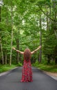 Vertical shot of a woman in a red dress standing on an empty road in a forest with happy open arms Royalty Free Stock Photo