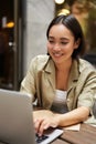 Vertical shot of young asian woman working on remote from outdoor cafe, sitting with laptop and smiling, studying Royalty Free Stock Photo