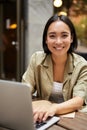 Vertical shot of young asian woman working on remote from outdoor cafe, sitting with laptop and smiling, studying Royalty Free Stock Photo