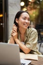 Vertical shot of young asian woman working on remote from outdoor cafe, sitting with laptop and smiling, studying Royalty Free Stock Photo