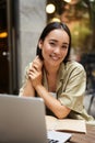 Vertical shot of young Asian woman working on remote from outdoor cafÃÂ©, sitting with laptop and smiling, studying Royalty Free Stock Photo