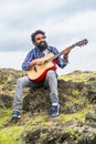 vertical shot of Young beard man playing guitar by singing song while sitting on top of hill - concept of hobbies Royalty Free Stock Photo