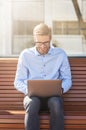 Vertical shot of a young happy businessman using laptop while sitting on the bench outdoors on a sunny day and working Royalty Free Stock Photo