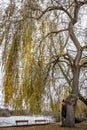 Vertical shot of young woman with mask looking at the camera leaning on willow tree on the lakeshore Royalty Free Stock Photo