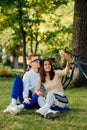 Vertical shot of a youthful smiling couple taking selfies on a smartphone and sitting on a green grass in the city park. Two