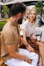 Vertical side view shot of elegant young woman with blond hair talking with bearded tattooed man sitting on bench with Royalty Free Stock Photo