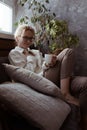 Vertical smiling, calm, glad blond woman in eyeglasses, in warm sweater sitting on sofa and drink hot tea. Coze weekend Royalty Free Stock Photo
