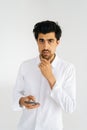 Vertical studio portrait of thinking young man in casual shirt looking at camera holding smartphone standing on white Royalty Free Stock Photo