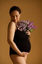 Delightful pregnant woman in black bodysuit, posing with purple lilacs on beige background, enjoying her pregnancy time Royalty Free Stock Photo