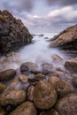 A vertical sunset seascape photograph of misty waves crashing on the rocks