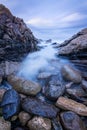 A vertical sunset seascape photograph of misty waves crashing on the rocks