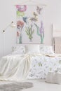 Vertical view of a scandinavian style bedroom interior with a bed dressed in white linen with painted green plants. Fabric wall ar