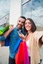 Vertical. Young caucasian real couple smiling and having fun taking a selfie portrait holding shopping bags at city