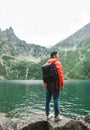 Vertical. Young tourist standing with his back on the shore of a beautiful lake in the rain on a background of mountains, wearing