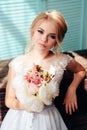 Very beautiful blonde with blue eyes in a white bride dress near a window with a bouquet of flowers Royalty Free Stock Photo