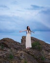 A beautiful girl in a white dress with dreadlocks is standing on a cliff with her arms outstretched against a cloudy sky at sunset Royalty Free Stock Photo