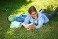 Very cute, young boy in round glasses and blue shirt reads book lying on the grass next to backpack and globe. Education Royalty Free Stock Photo