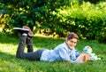 Very cute, young boy in round glasses and blue shirt reads book lying on the grass next to backpack and globe. Education Royalty Free Stock Photo