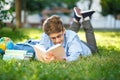 Very cute, young boy in round glasses and blue shirt reads book lying on the grass next to backpack and globe. Education Royalty Free Stock Photo