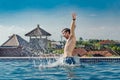 Very happy, slender young smiling man, jumping out of pool water with splashes, on roof, and hands up.