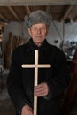 Very old man in retro clothes stands among his workshop at home, holds wooden cross in his hands and looking at the Royalty Free Stock Photo