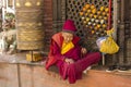 A very old Tibetan Buddhist monk in red and yellow clothes is sitting near a rotating prayer drum