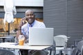 A very smiling black entrepreneur talking on the phone and working remotely with his laptop. He is sitting on the terrace of a