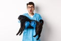 Vet clinic concept. Amazed male doctor veterinarian holding cute black pug dog, smiling and staring up impressed Royalty Free Stock Photo