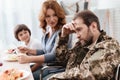 A veteran in a wheelchair dines with his family. A man in uniform is sitting at the kitchen table.