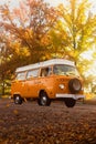 Vibrant orange camper van is parked in an autumnal setting, surrounded by trees in full foliage