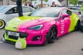 A vibrant, pink and neon green sports car sits on a public street surrounded by other vehicles and pedestrians The cars