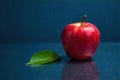 A vibrant red apple rests on a smooth, dark blue surface, accompanied by a single, fresh green leaf reflecting in the polished Royalty Free Stock Photo